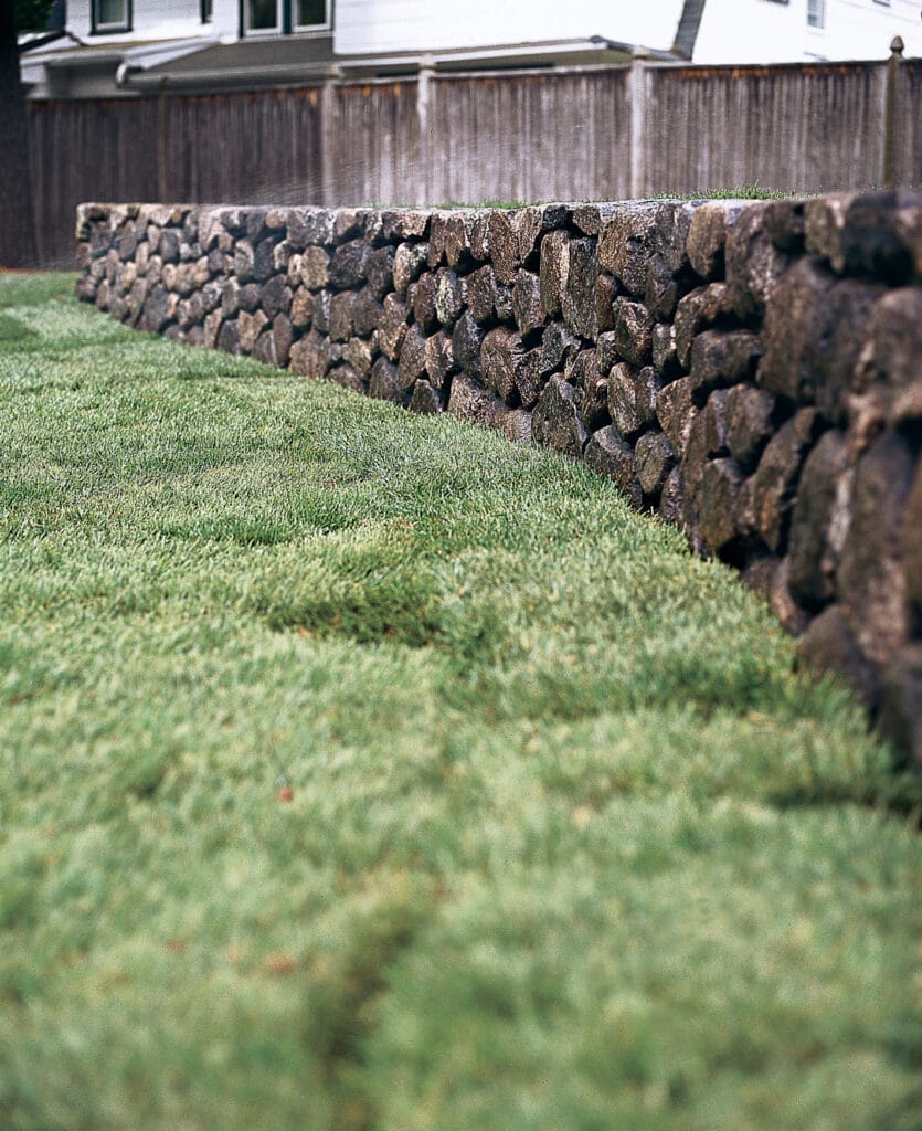 A neatly laid sod lawn extends up to a stone retaining wall, with a wooden fence and house in the background.