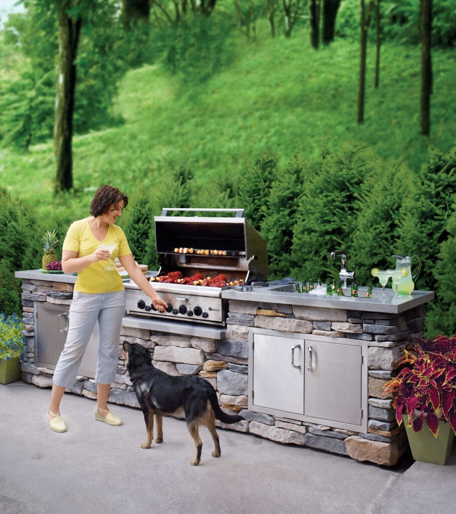 A woman standing in front of an outdoor kitchen with a grill and sink. A dog stands in front of her.