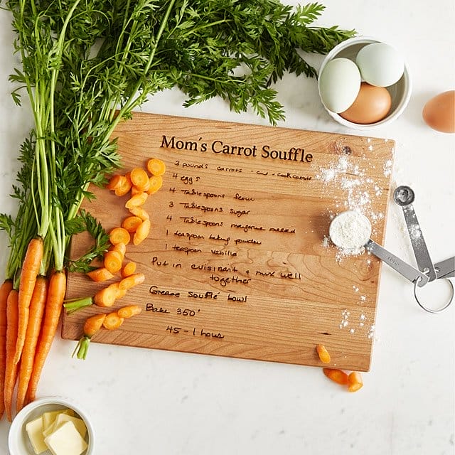 A cutting board with a recipe titled "Mom's Carrot Souffle", covered in carrots, flour, and eggs.