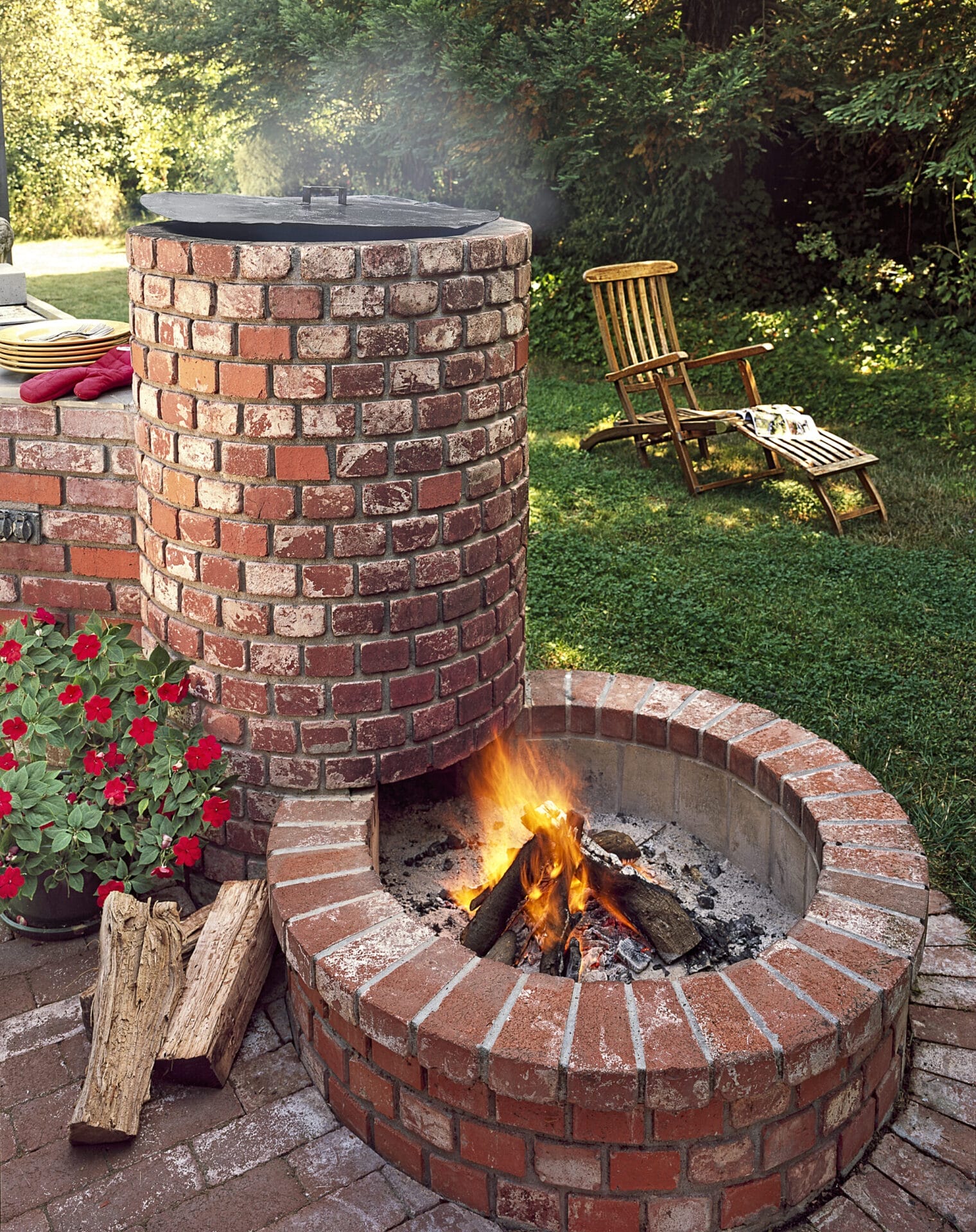 A homemade ted brick firepit beside a plant with red flowers and a wooden chair.