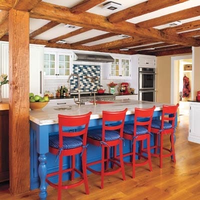 Renovated kitchen with white cabinets, marble countertops, exposed beams, and red-white-blue island maintaining colonial character.