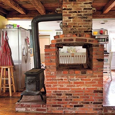 Historic kitchen before renovation featuring rustic hand-hewn ceiling beams and brick chimney.