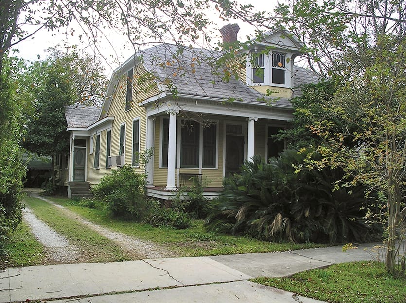 Overgrown yellow Creole cottage with two front doors partially hidden by wild vegetation.