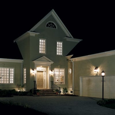 A garage and driveway lit up by three lights mounted on the house and one post lantern.