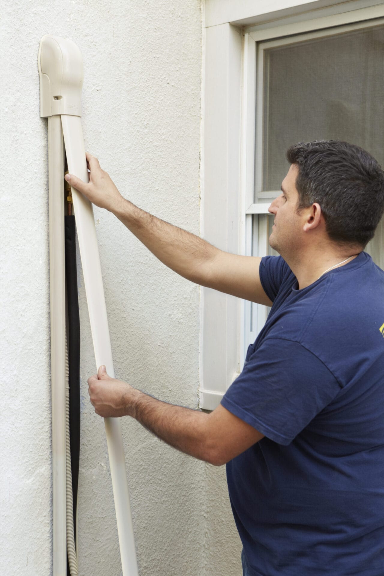 A man affixing a plastic channel to the siding of a house to hide AC lines.