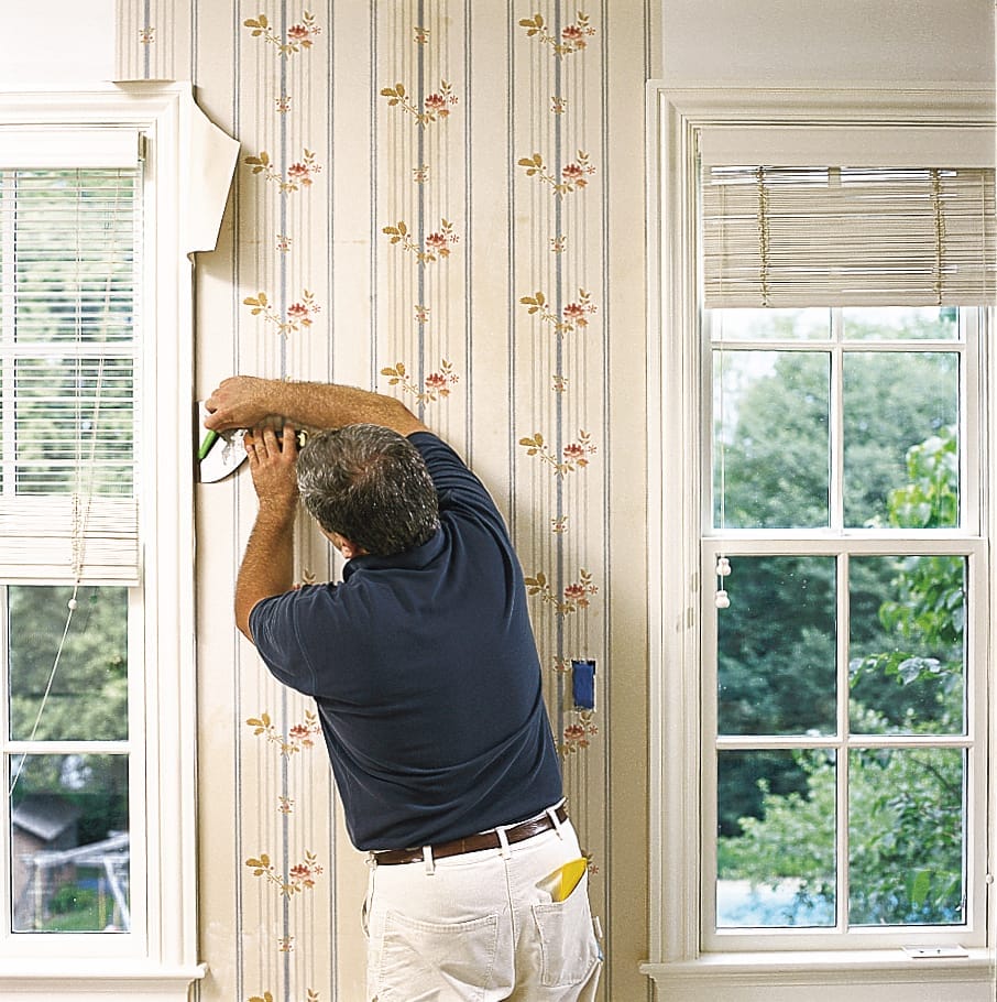A man trimming newly hung wallpaper around the windows and doors of the building.