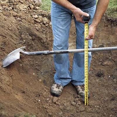 Person digging hole with shovel while measuring depth with measuring tape.
