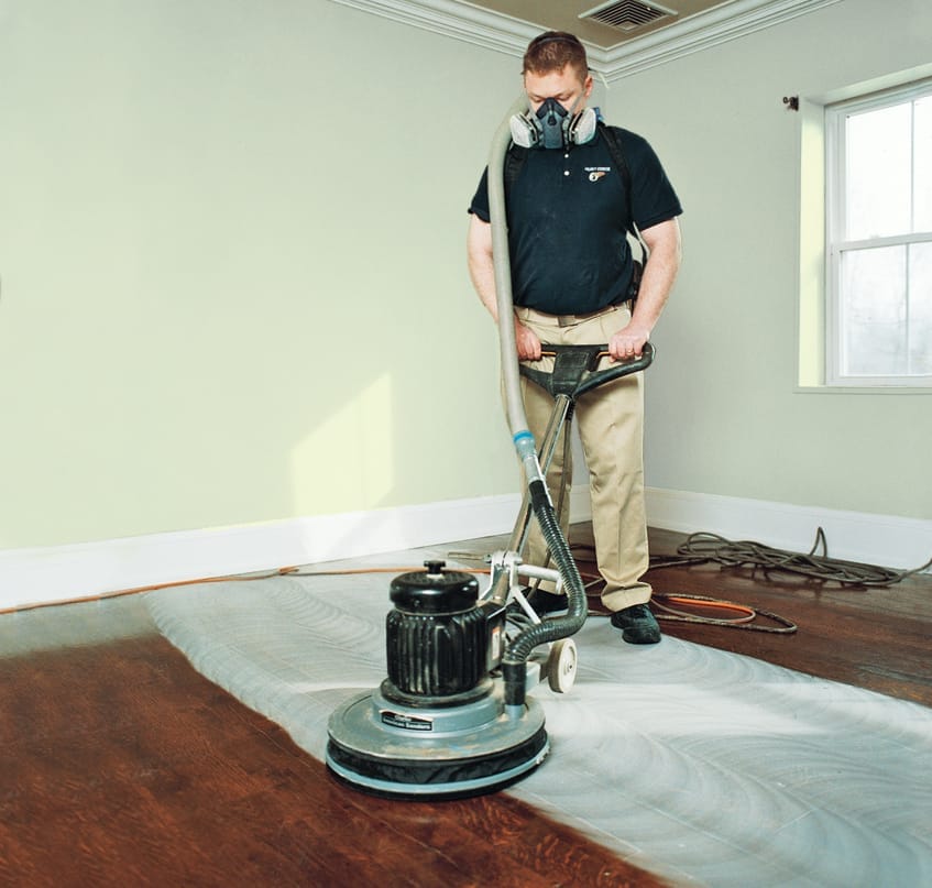 A man wearing a dust mask uses a floor buffer to scuff-sand the floor for finishing.