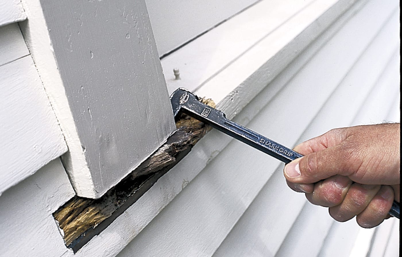A person uses a prybar to remove rot damaged wood from a house's siding.