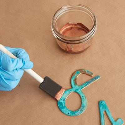 Image of a person adding more copper surfacer to metal numbers as the fourth step in creating a copper patina finish
