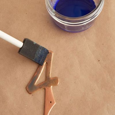 Image of a person adding more blue patina solution to metal numbers as the third step in creating a copper patina finish