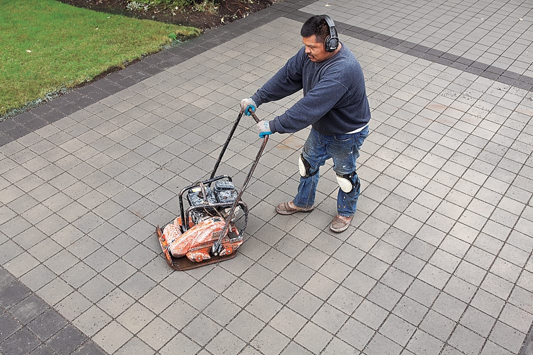 Man runs a plate compactor over the surface of the permeable paver driveway.