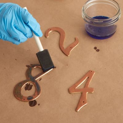 Image of a person adding a blue patina solution to metal numbers as the second step in creating a copper patina finish