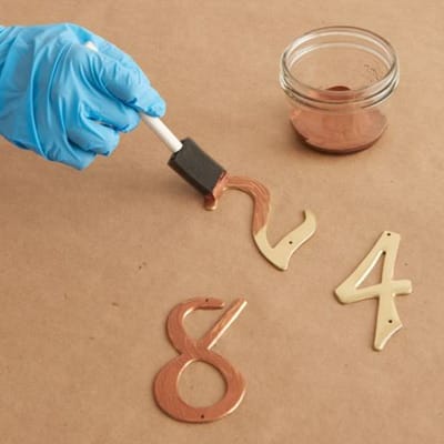 Image of a person adding copper surfacer to metal numbers as the first step in creating a copper patina finish