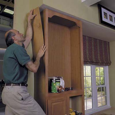 A man using a board of wood to conceal the joints of a recently installed book case.