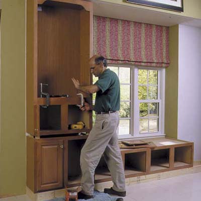 A man fitting a bookcase on top of a cabinet beside a window.