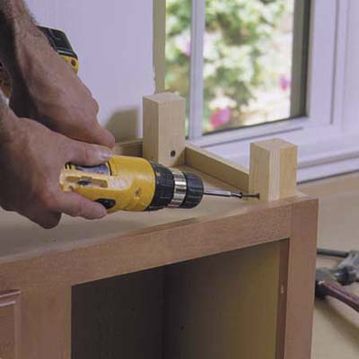 A man using a drill to fasten wooden blocks to the top of a cabinet.