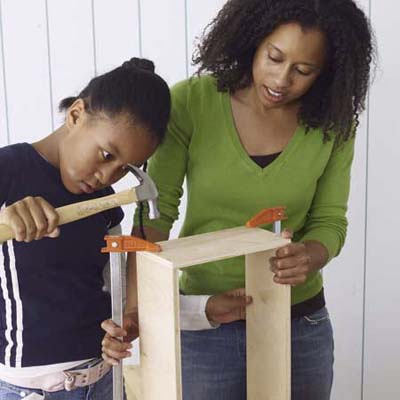 A woman holds pieces together while a child uses a hammer to put together a toolbox.