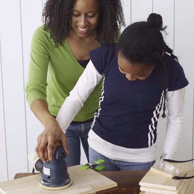 A woman helps a child sand wood for a toolbox.