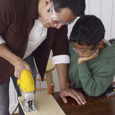 A child watches a man cut a handle out for a toolbox.