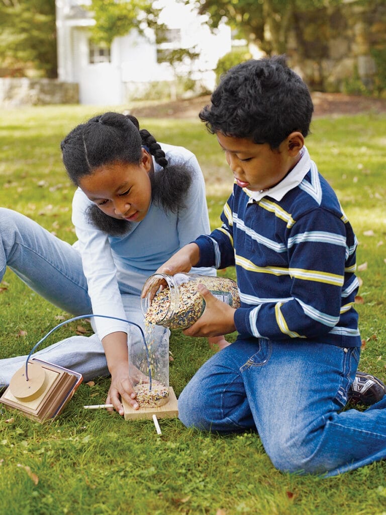 Two children pouring bird seed into a feeder.
