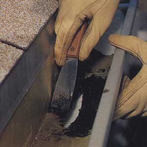 Person with gloved hands applying roofing cement around a hole in a gutter.