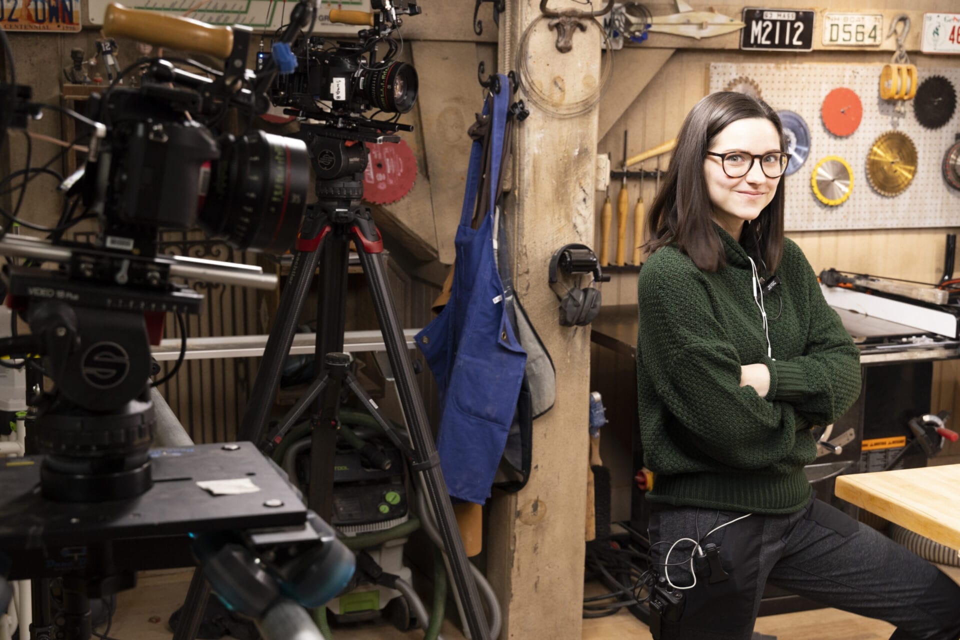Sarah Chasse, wearing glasses and a green sweater, stands confidently with arms crossed in a workshop filled with tools, equipment, and cameras. She is ready for filming, with a camera tripod set up in the foreground.