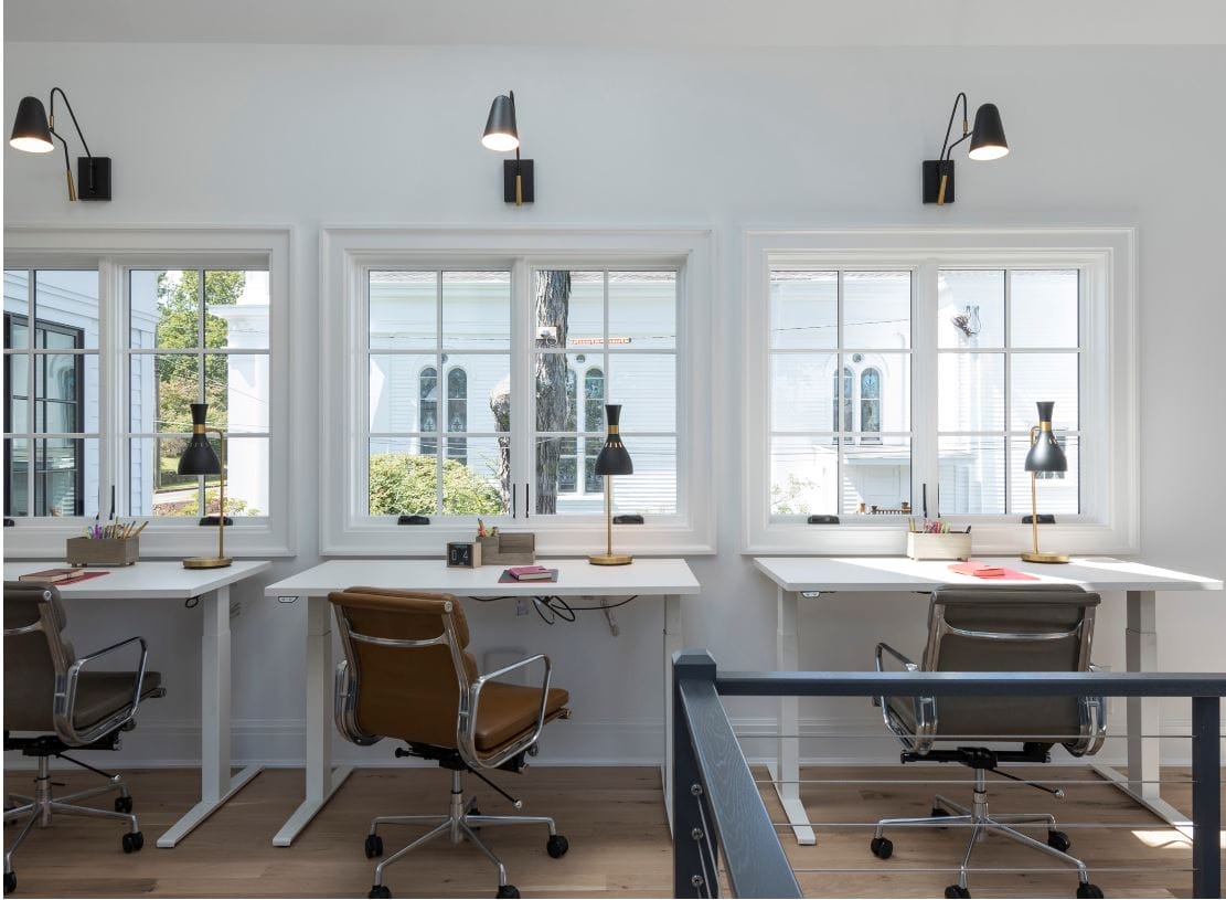 Row of adjustable height desks under wide windows overlooking neighboring church with wall sconces and table lamps.
