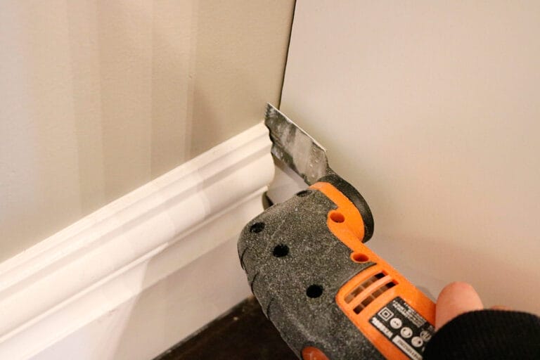 An oscillating tool with a flat blade being used to trim and remove a section of white baseboard trim along a wall where the built-in Ikea bookcase will go.