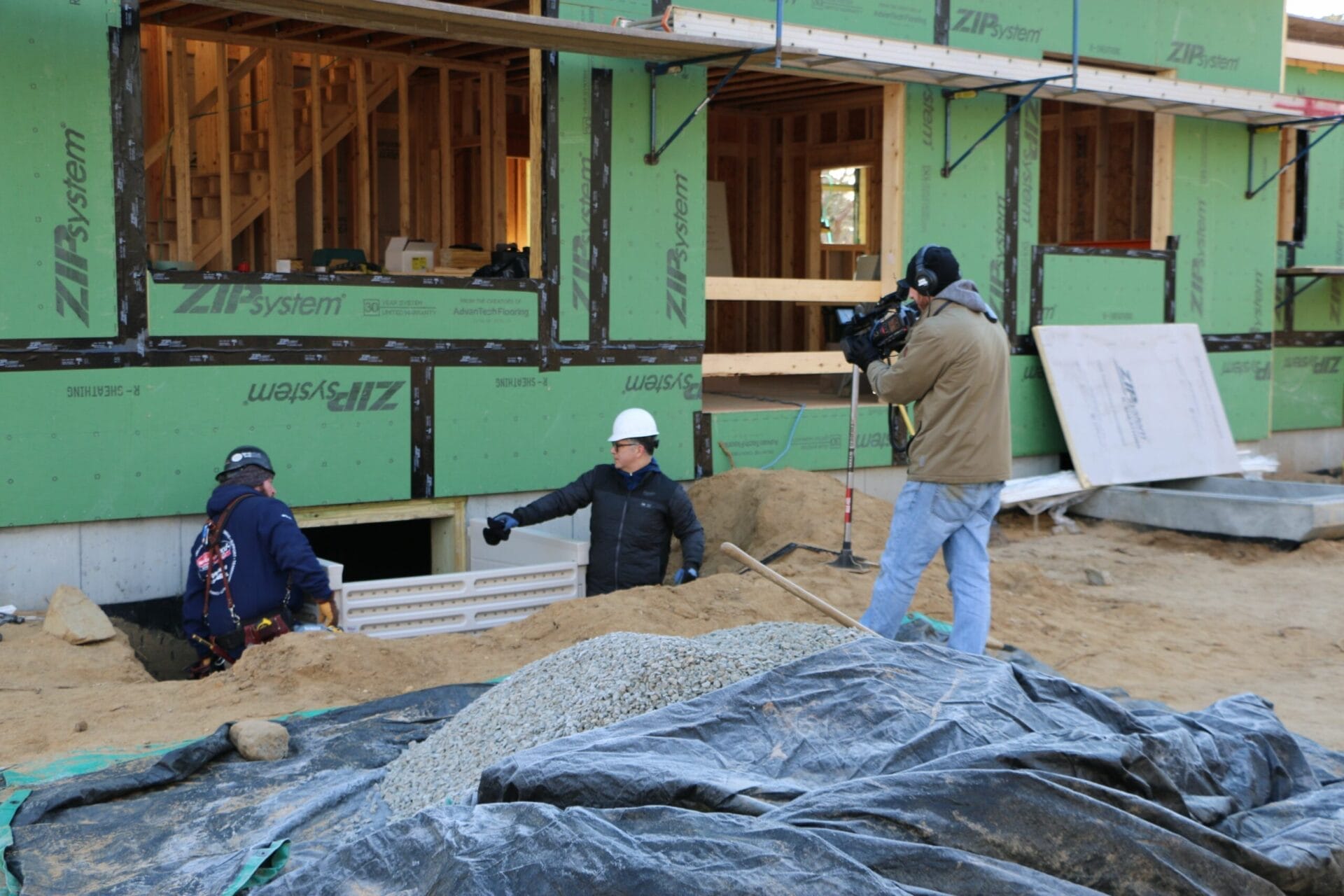 A cameraman films two men discussing the installation of an egress window well.