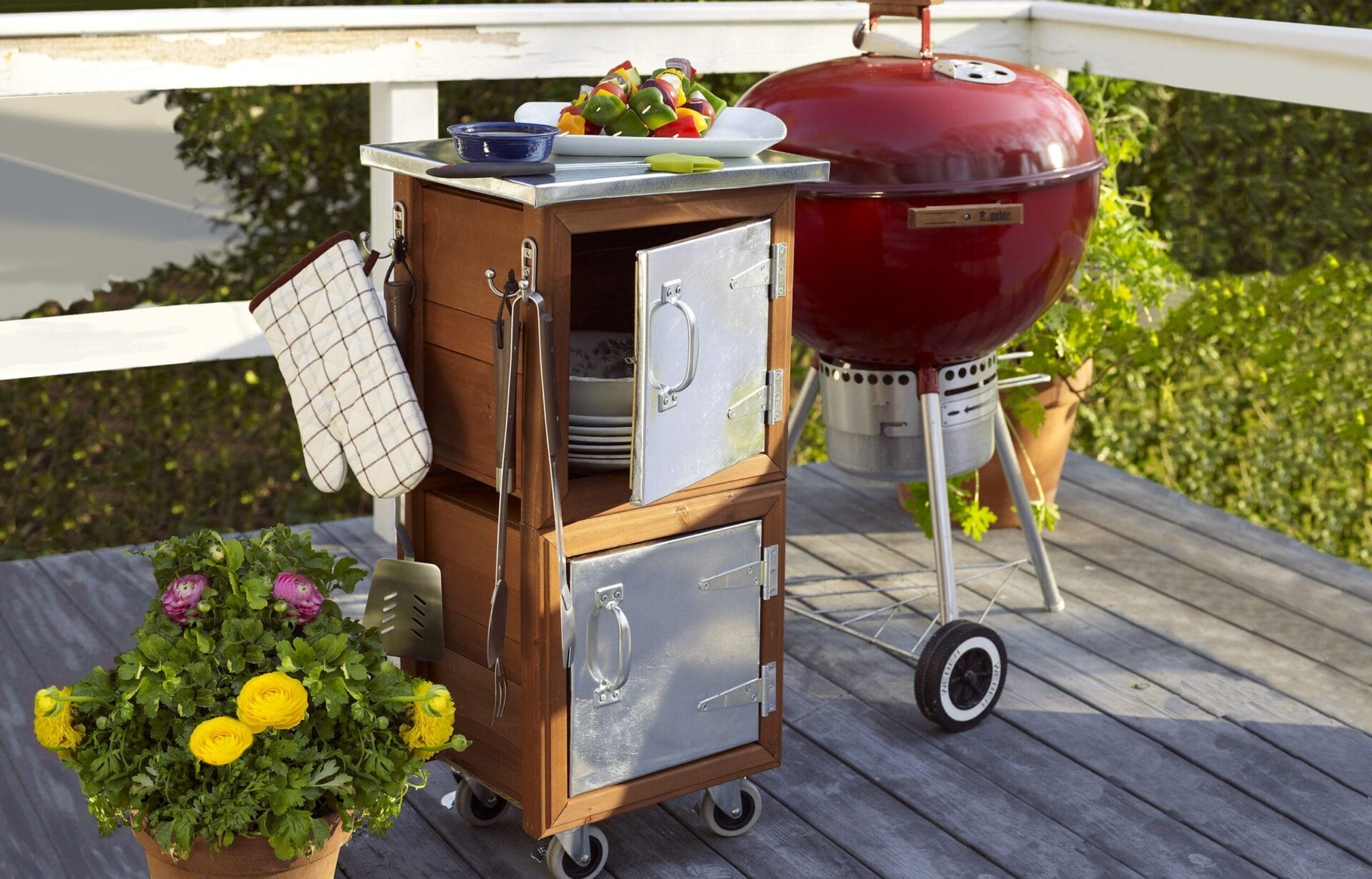 A red charcoal grill and a wooden rolling cart with metal doors, perfect accessories you need if you love to grill, sit on a wooden deck. The cart holds plates, utensils, and vibrant vegetables. Potted plants with flowers decorate the deck against a lush green backdrop.