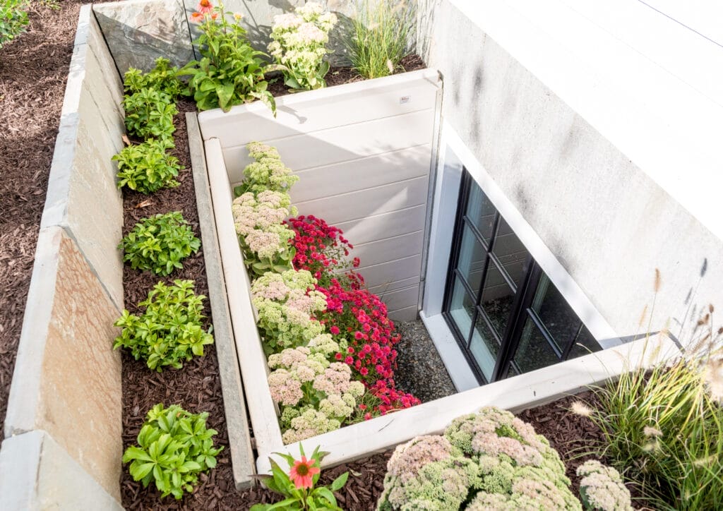 A closed egress window surrounded by plants and flowers.