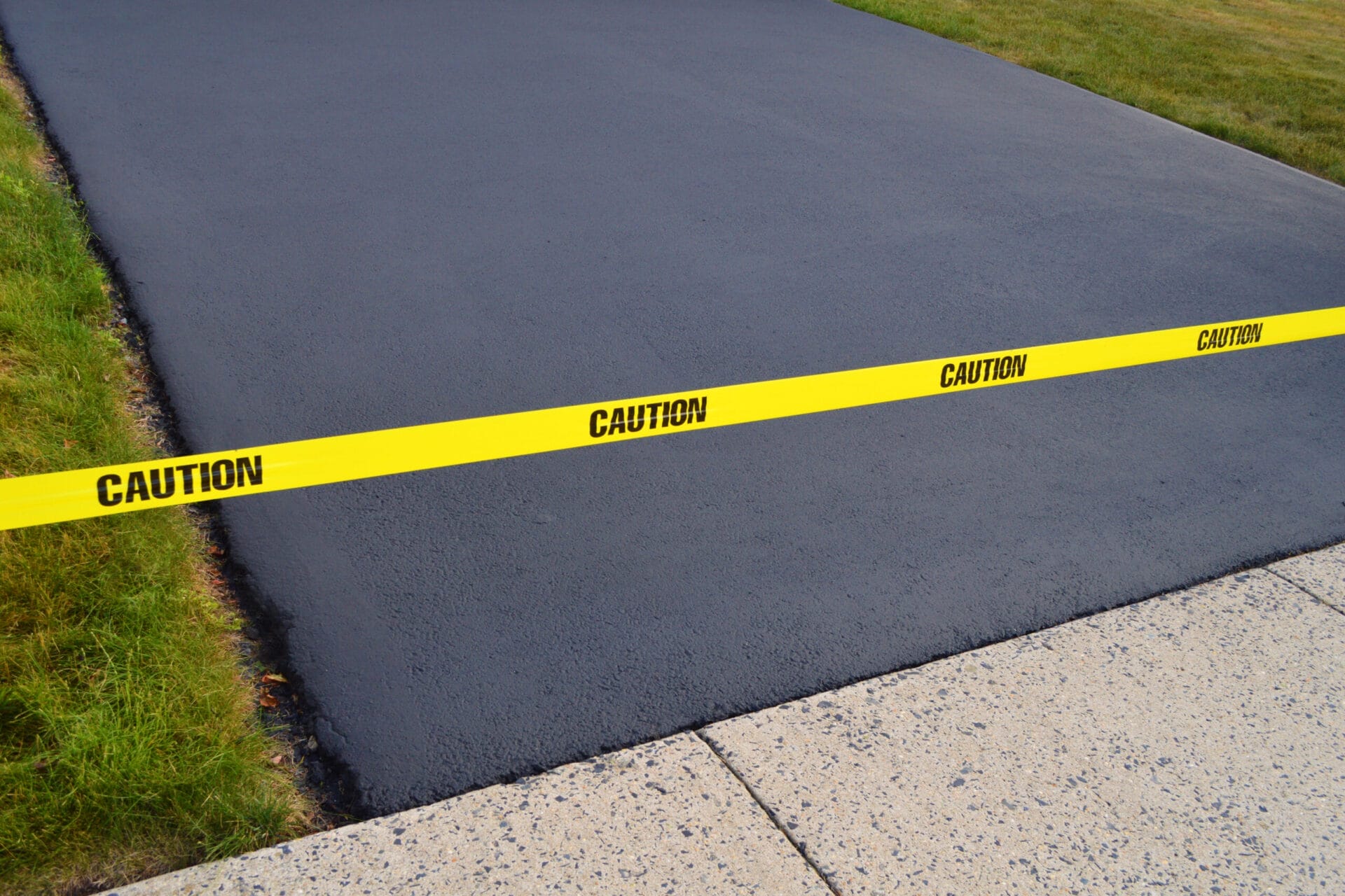 A freshly paved driveway with pristine sealcoat is marked with yellow caution tape. Lush green grass borders the sleek black asphalt on one side, while a light-colored concrete sidewalk runs along the bottom.
