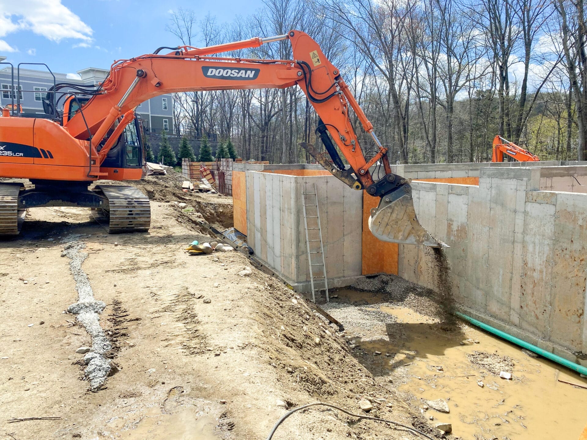Excavator placing gravel around drainage pipes at foundation bottom.