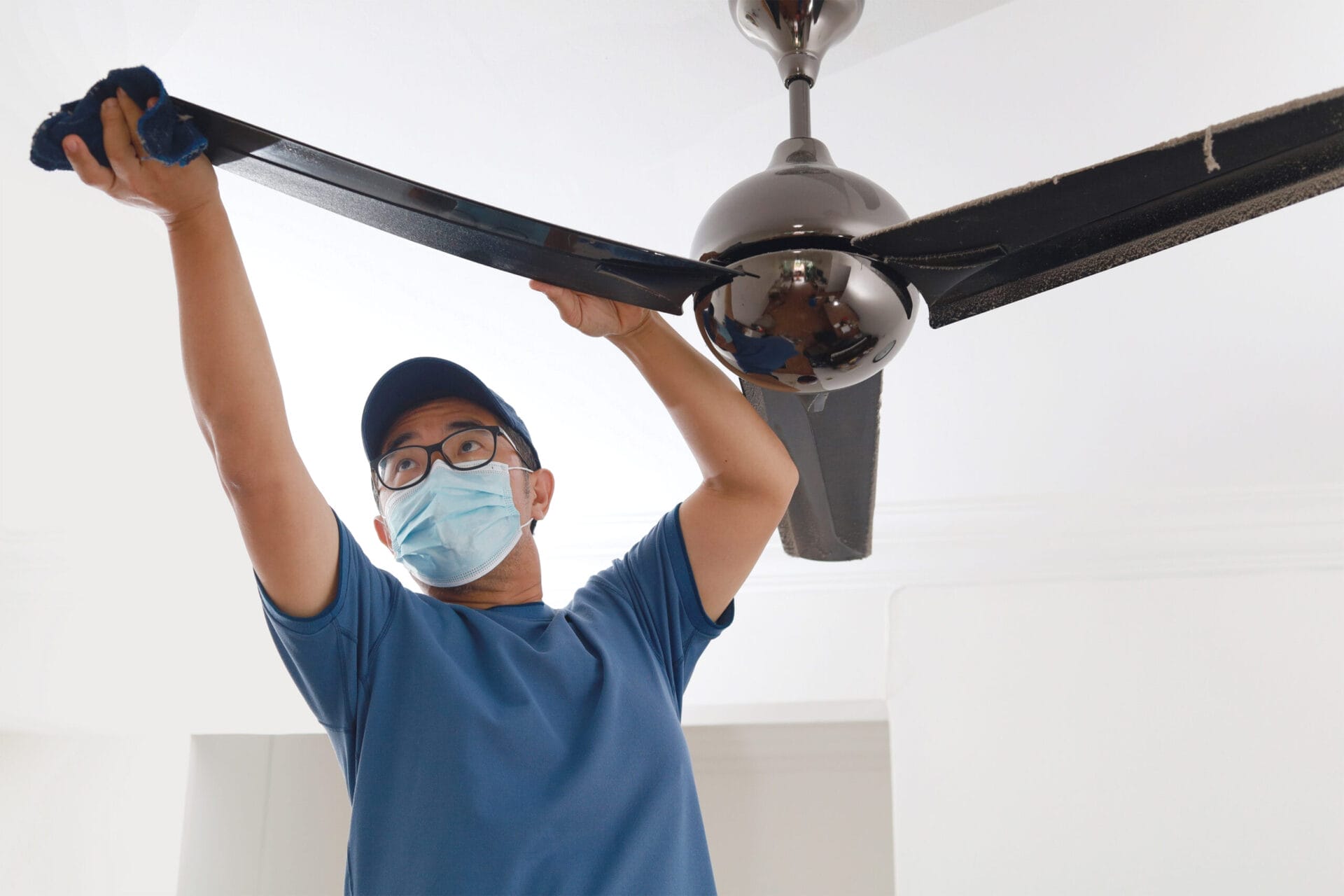 A man cleans a ceiling fan in his home.