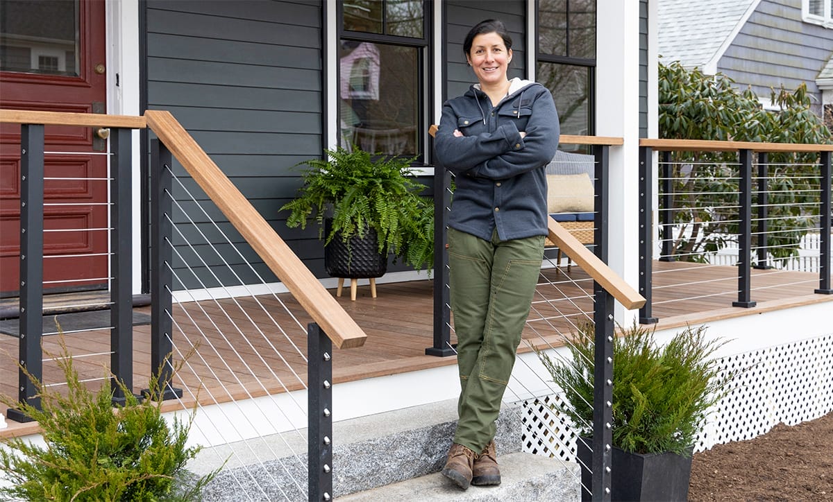 Landscape Contractor Jenn Nawada stands on the steps of a porch, arms crossed and smiling. They are wearing a dark jacket, green pants, and brown boots. The porch has cable railing with wooden handrails and potted plants beside the steps.