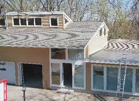 The front exterior of this Lexington Modern two-story house boasts beige siding and a complex roof structure with multiple gables and a dormer. Large windows, an open garage door on the left, an open front door, and a closed sliding door on the right side create a welcoming facade. A ladder rests on the right side amidst leafless trees in the background.