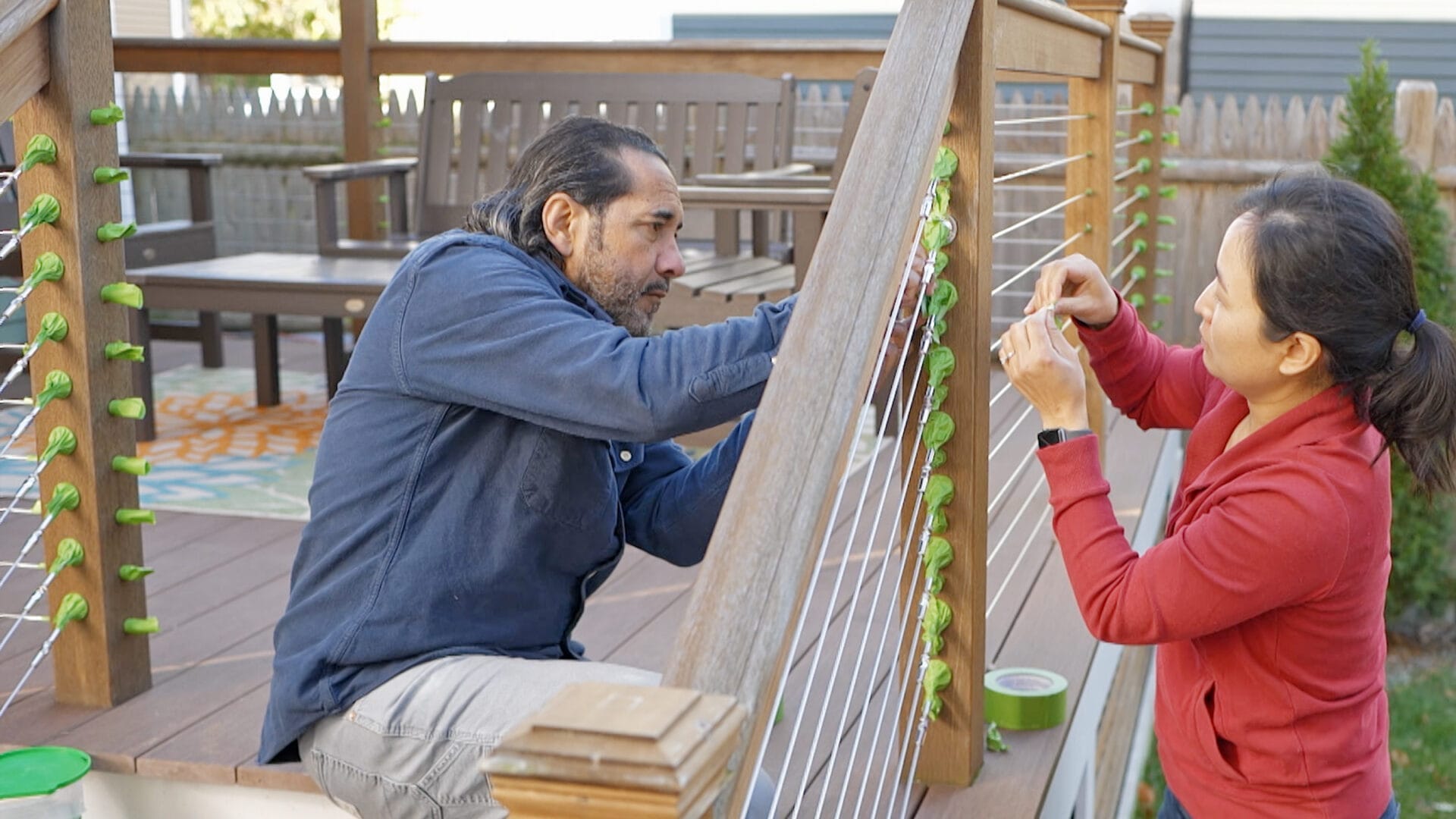 Two people are working together on a deck. The person on the left kneels, securing string lights to a wooden railing, similar to prepping for paint. The person on the right holds the end of the lights. A patio area with chairs is visible in the background, ready for evening enjoyment.