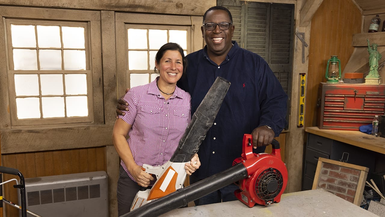 Two people smiling in a workshop. One holds a leaf blower while the other drapes an arm around them.