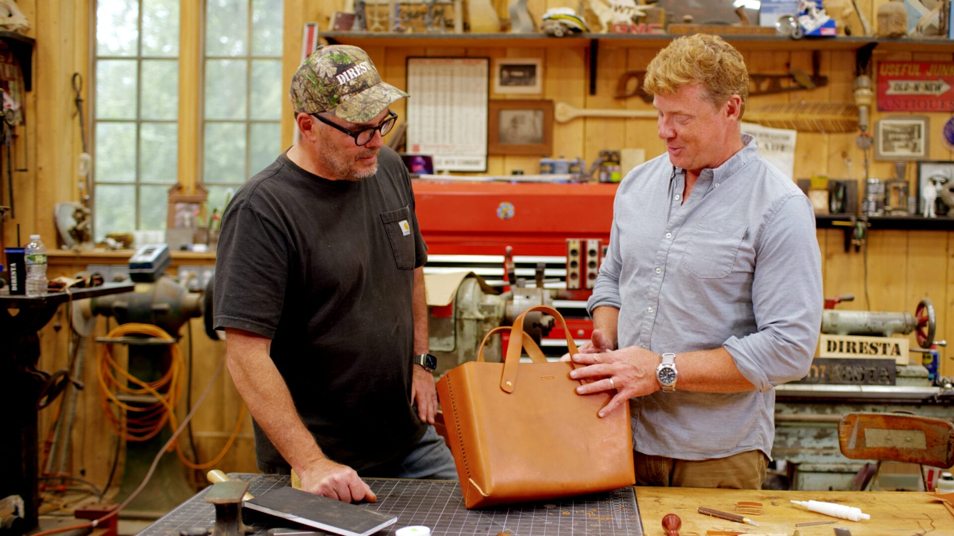 Two men stand in a workshop. One wears a cap and T-shirt; the other wears a light blue shirt. Tools and machinery are visible in the background.