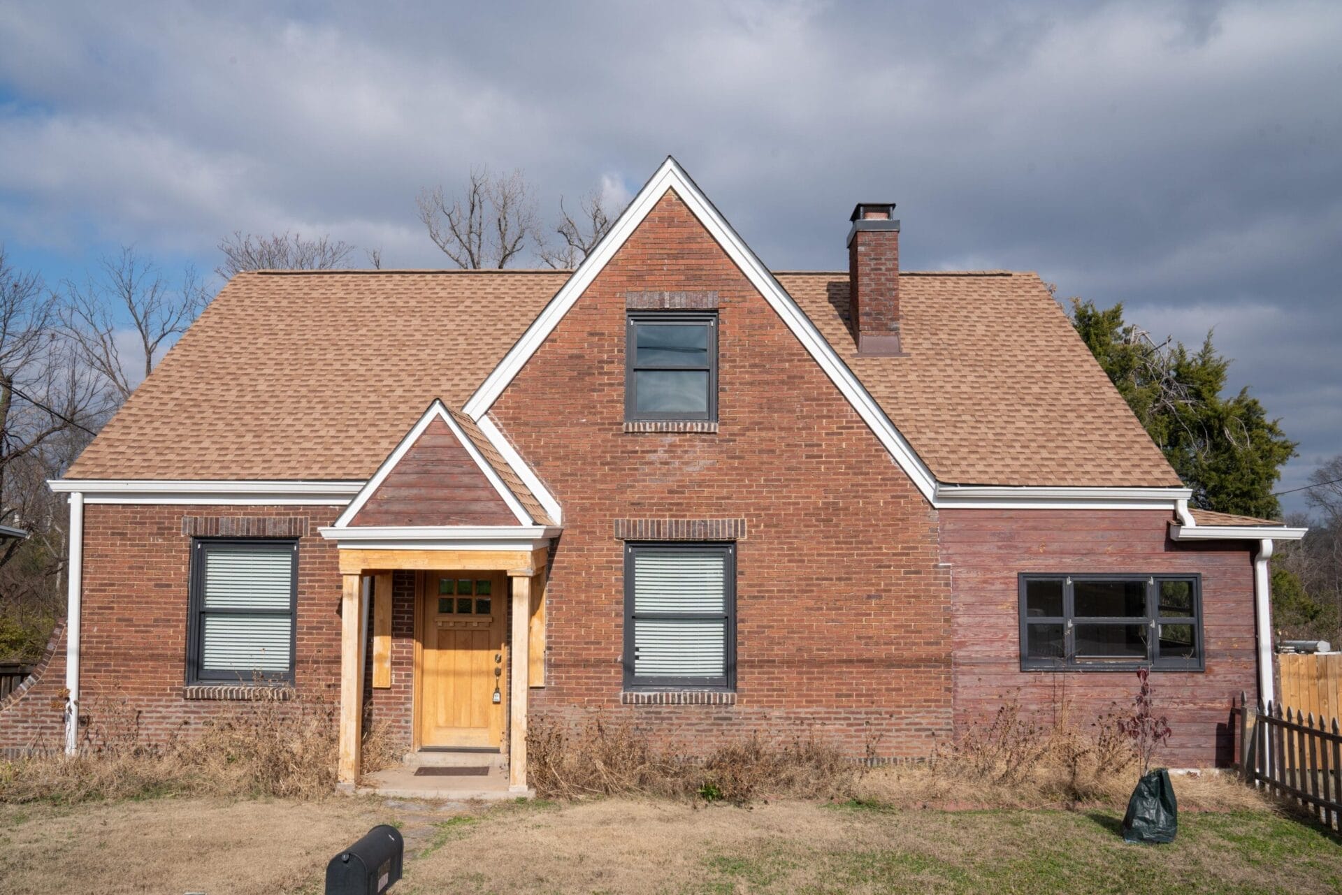 A brick house with a steep, triangular roof and a contrasting light brown shingled section. It features a wooden front door, two windows with shutters, and a chimney. The front yard is grassy with a mailbox and sparse vegetation. Cloudy sky in the background.
