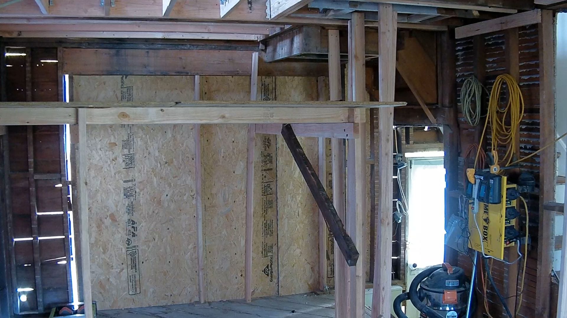 The partially constructed interior room, part of a Ridgewood Colonial Revival, features exposed wooden framing and a plywood wall. Tools and cables are visible in the corner as sunlight streams through a window, illuminating the space like a scene captured on a kitchen webcam.