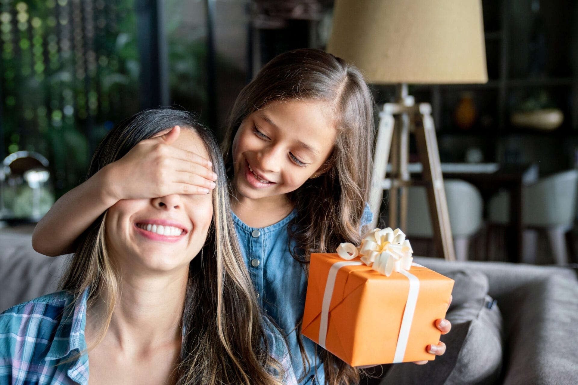 A smiling young girl, holding a box wrapped in orange paper with a white ribbon and bow, covers her mother's eyes to surprise her
