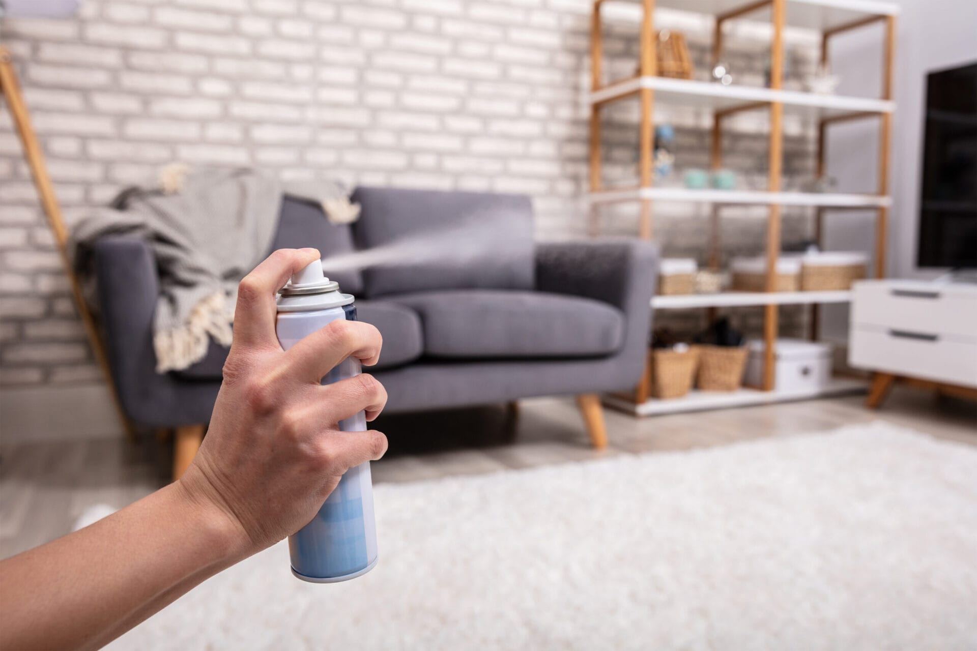 A hand spraying air freshener in a living room exudes a sense of serenity. The house smells fresh amid a gray couch with a blanket, a white and gray rug, a brick wall, and a wooden shelf adorned with baskets and decorative items.